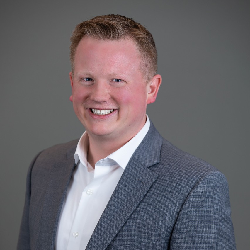 Josh, wearing a white dress shirt and gray suit jacket, smiles at the camera.