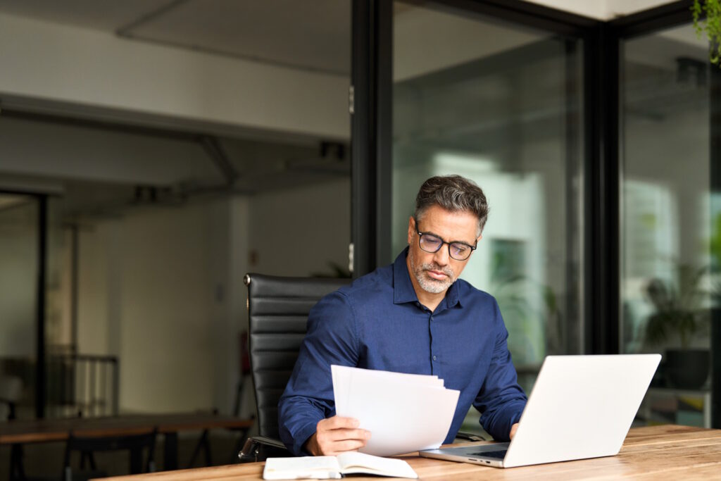 A man is sitting at a desk in a glass office looking through papers with his laptop open in front of him.