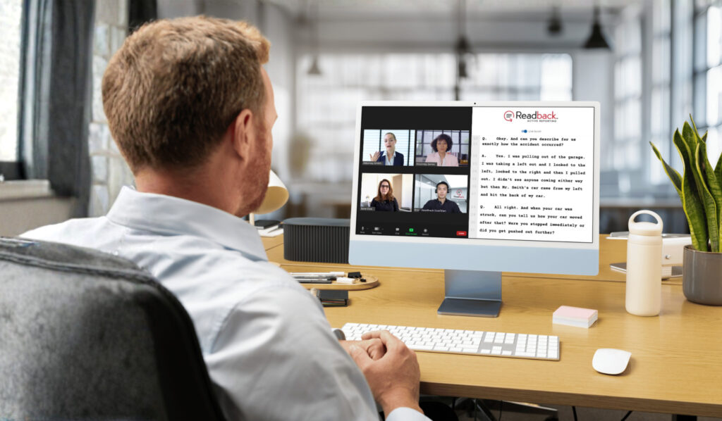 A man with short blonde hair viewed from behind, wearing a blue shirt, sits in a dark chair at a light wood desk, looking at an all-in-one computer screen. The screen is split: the left side shows a video conference with four people in a grid, and the right side displays the "Readback" logo and transcribed text of a conversation. His hands are near a white keyboard and mouse. A white water bottle, plant, and sticky notes are on the desk. The right background shows a modern, open-plan office with desks and windows.